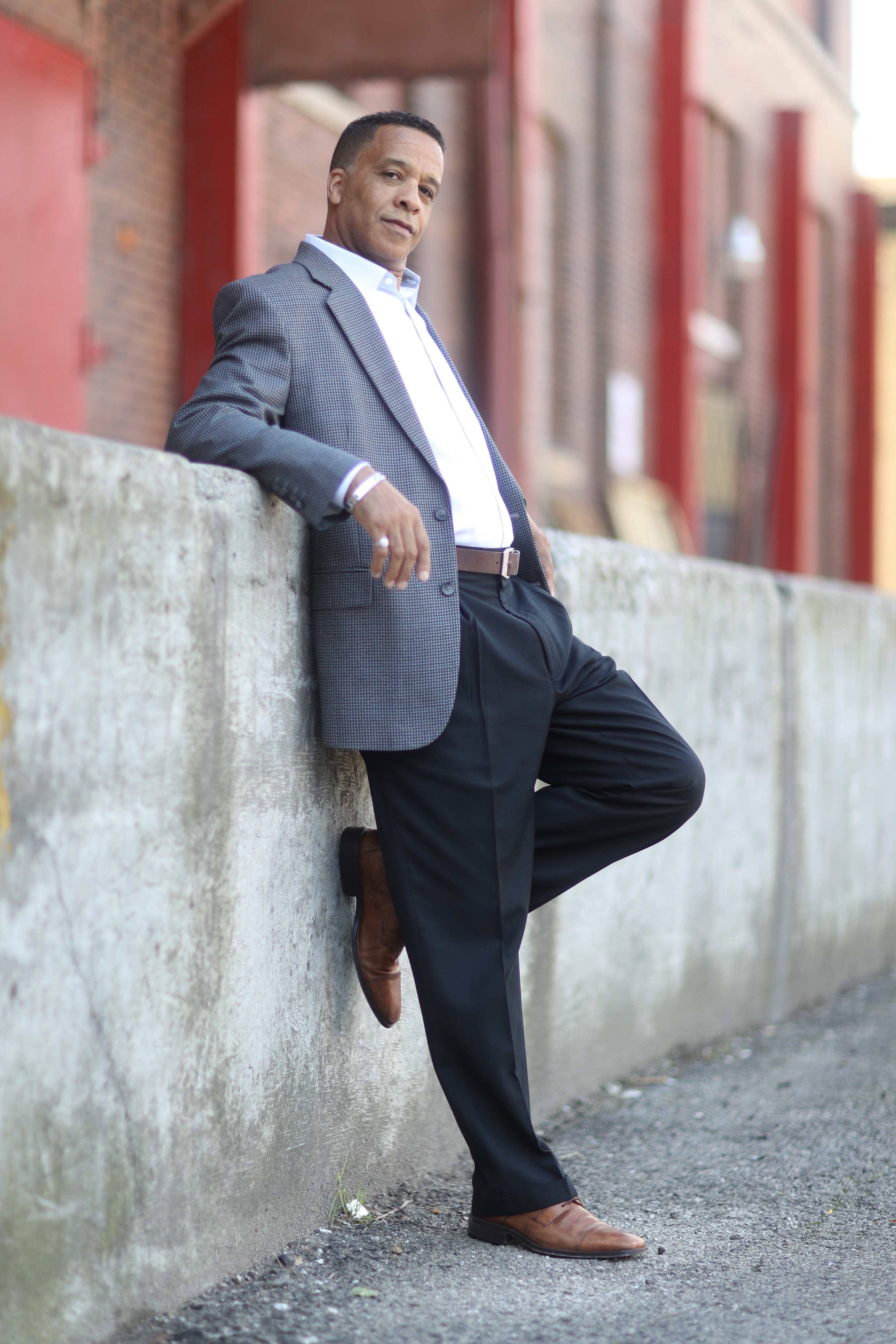 The image is the section that states, “What’s holding you back?” is of a well-dressed light brown skin-toned man leaning back against a concrete wall. Dark blue dress pants, brown belt, medium blue tweed jacket, white collared shirt, the top button was undone, no tie. he is facing to the right of the image and head turned to the camera. The left/ rear leg in the image is raised with the shoe against the wall at about knee height.